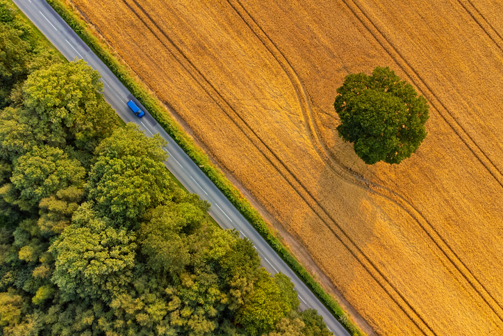 Wide angle aerial view of late summer fields and deciduous trees, and blue vehicle driving along country road.